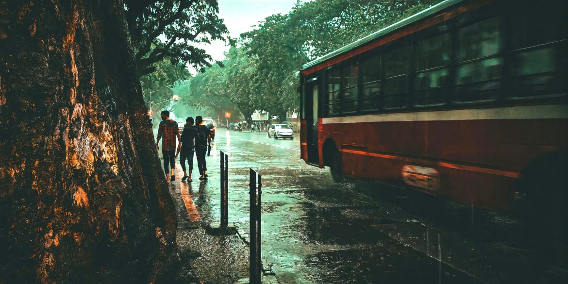 A group of people walking down a sidewalk next to a bus as it rains in Mumbai. Photo by Sadiq Sonalkar/Unsplash.